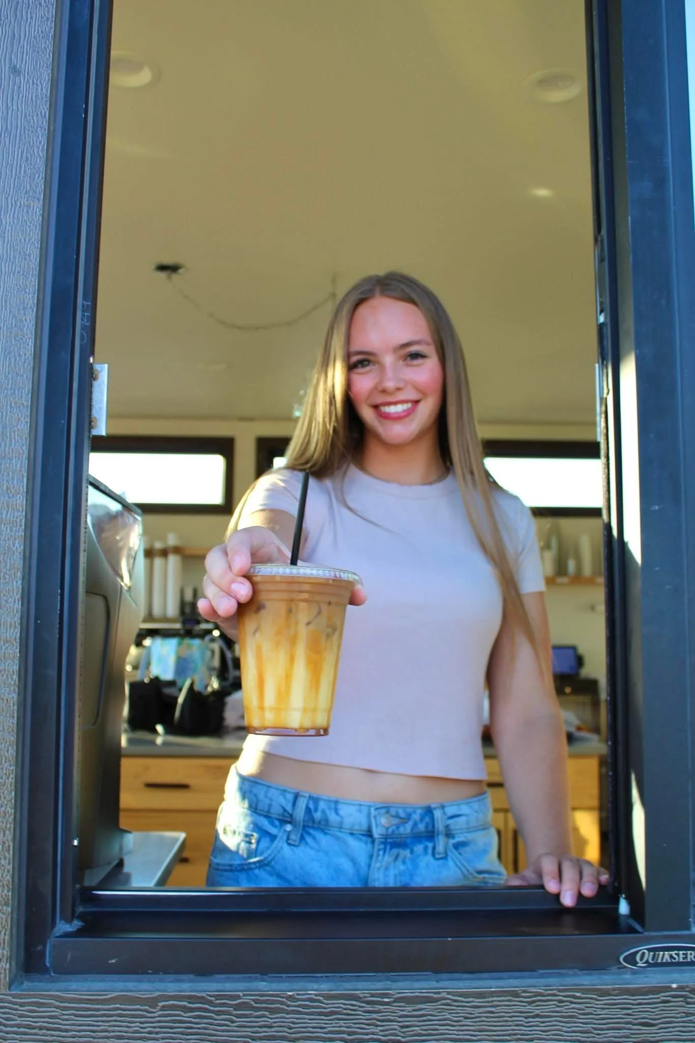 A young woman with long blonde hair, wearing a white t-shirt and denim shorts, is smiling as she takes a drink from a clear plastic cup with a black straw through a drive-thru window.