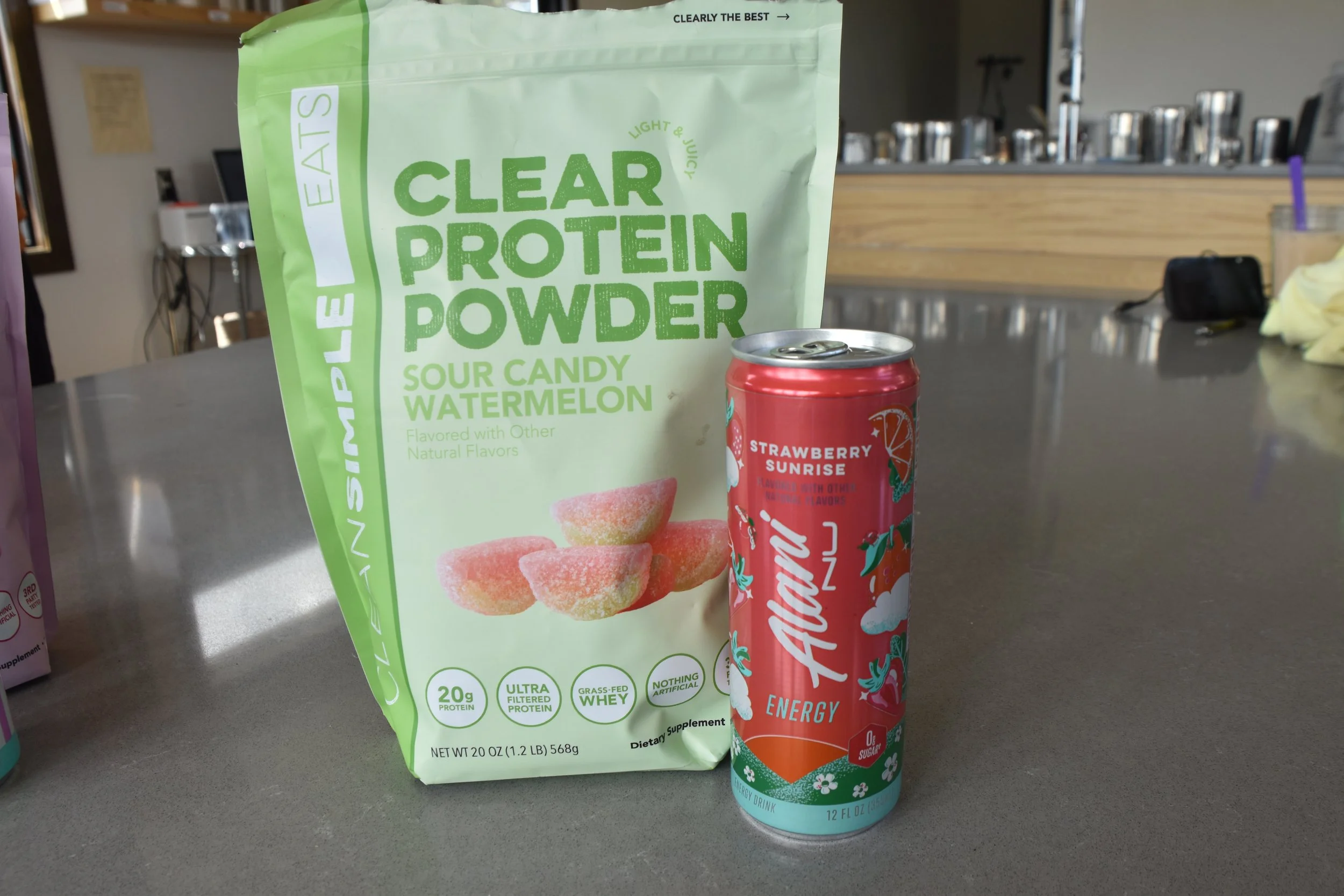 A green bag of Clear Protein Powder with flavor sour candy watermelon and an energy drink can labeled Aston Energy with strawberry flavor, both placed on a gray table in a room with a wooden counter and stainless steel containers in the background.