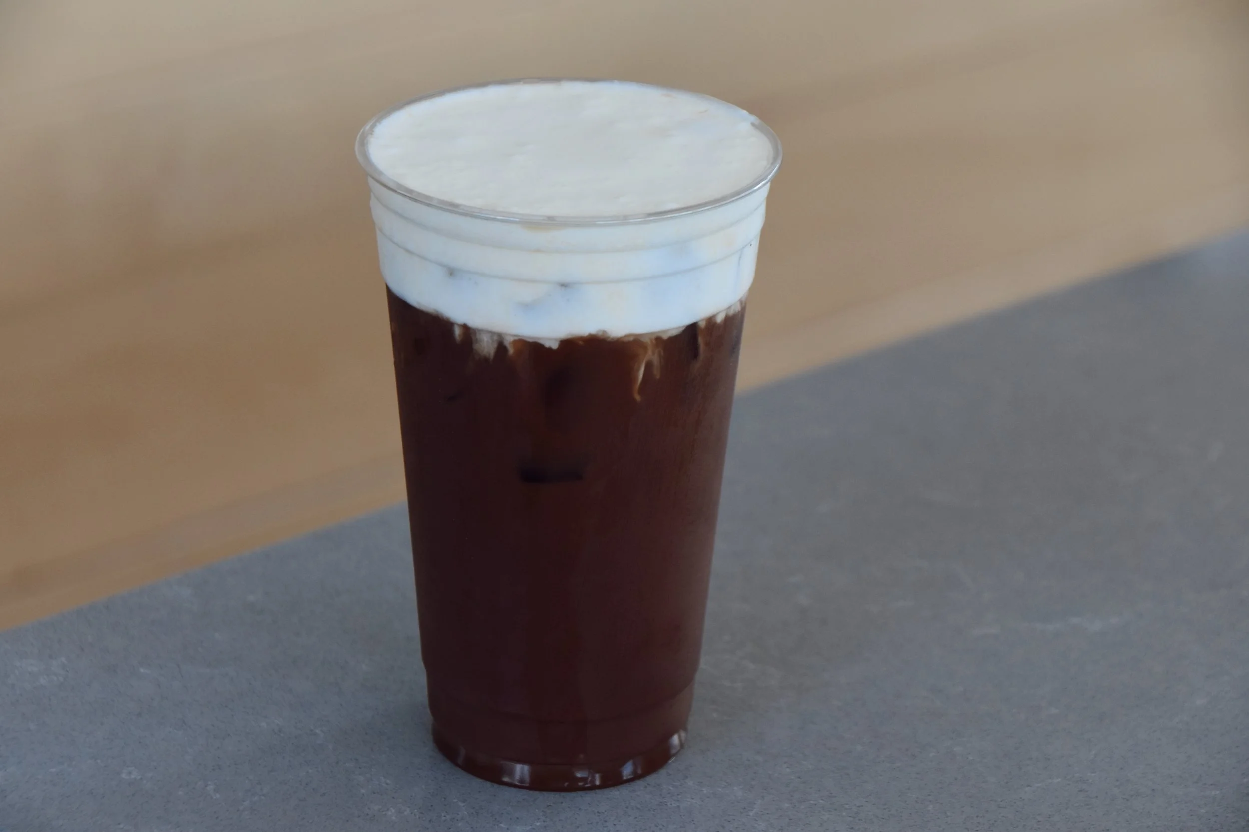 A clear plastic cup filled with iced coffee topped with whipped cream and chocolate syrup, sitting on a gray surface.