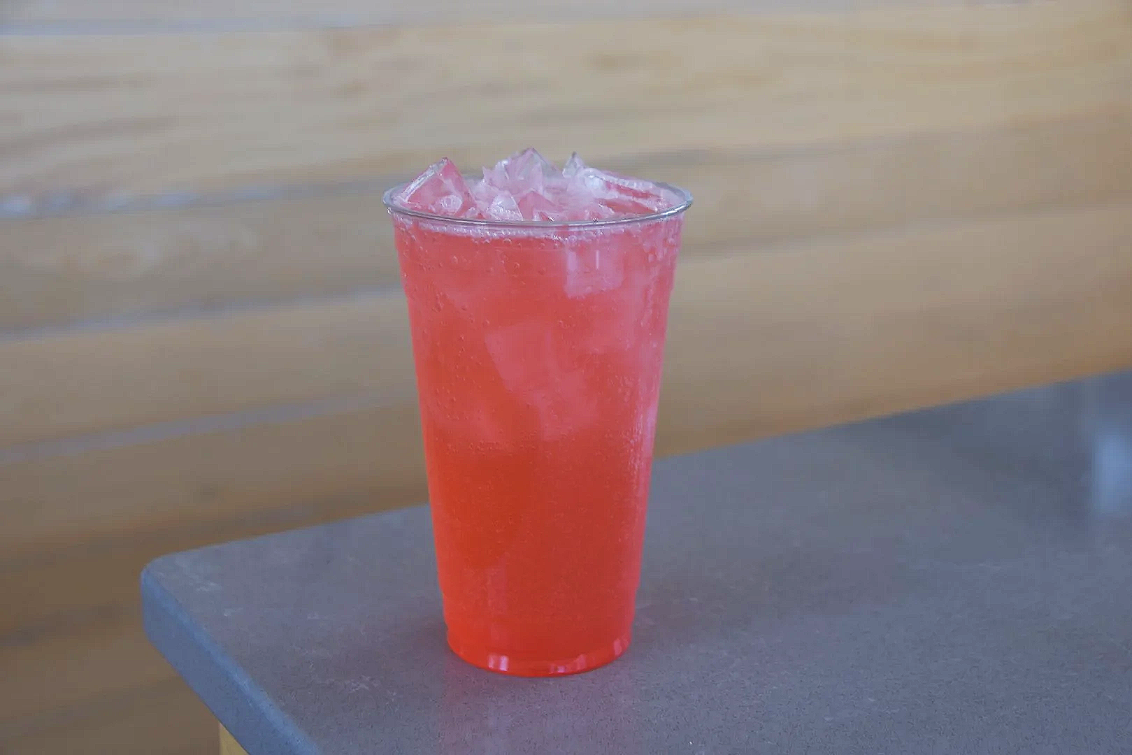 A clear glass filled with a bright pink, slushy icy drink topped with ice cubes, sitting on a gray surface with a wooden background.