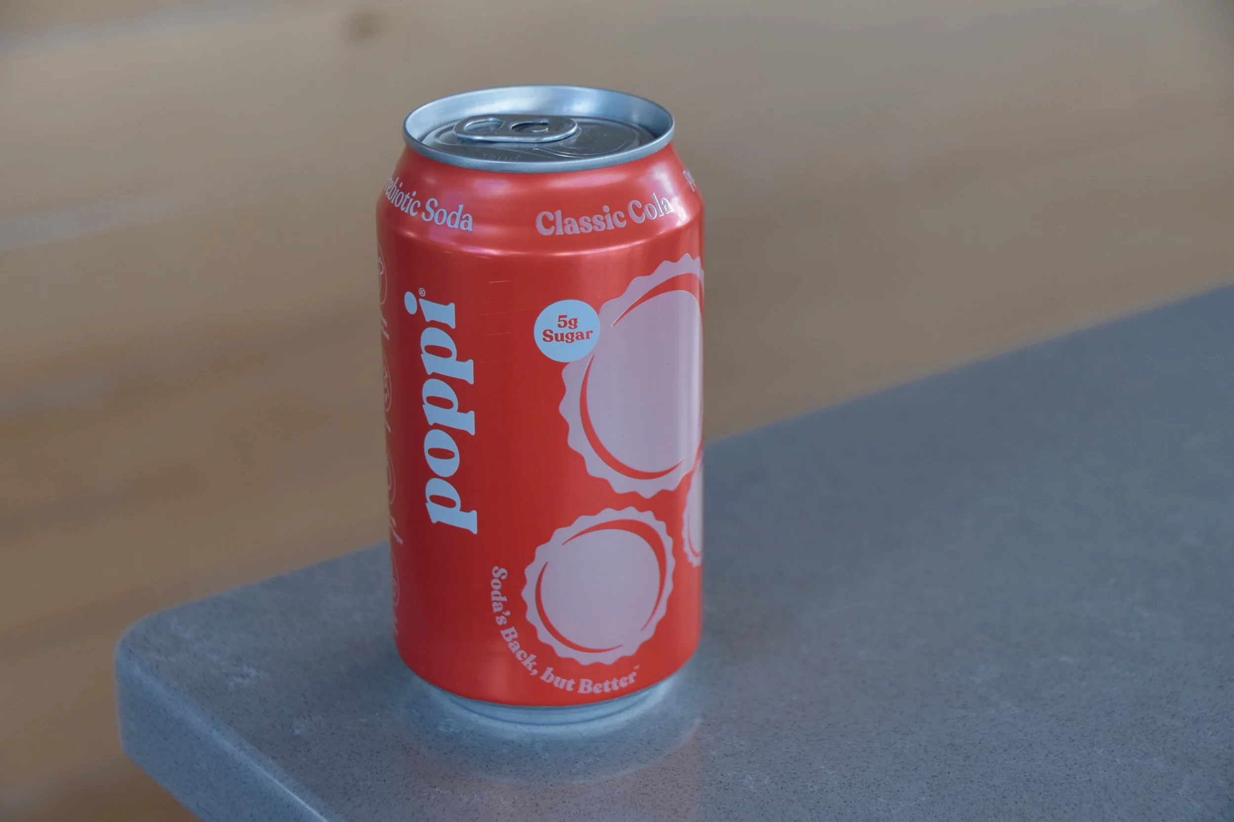 A red soda can placed on a blue cylindrical surface. The can is labeled with phrases like 'Probiotic Soda', 'Classic Cola', and notes '5g Sugar'. The background is out of focus.