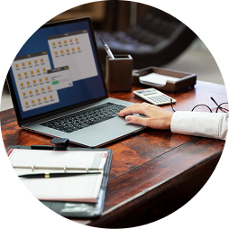 A person working on a laptop at a wooden desk with a calculator, notepad, and pen.
