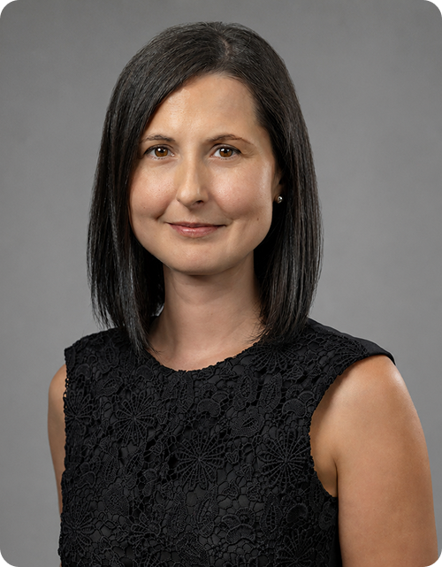 Professional woman with straight, shoulder-length dark hair, wearing a black lace sleeveless top, standing against a gray background.