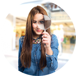 Young woman with long brown hair holding a magnifying glass, inspecting something closely in a bright indoor space.