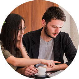 A man and woman sitting at a table, looking at a tablet together. The woman is smiling, and the man is focused on the device. There is a cup of coffee on the table.