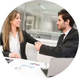 Two business professionals, a woman and a man, shaking hands in a modern office setting with documents and a laptop on the table.