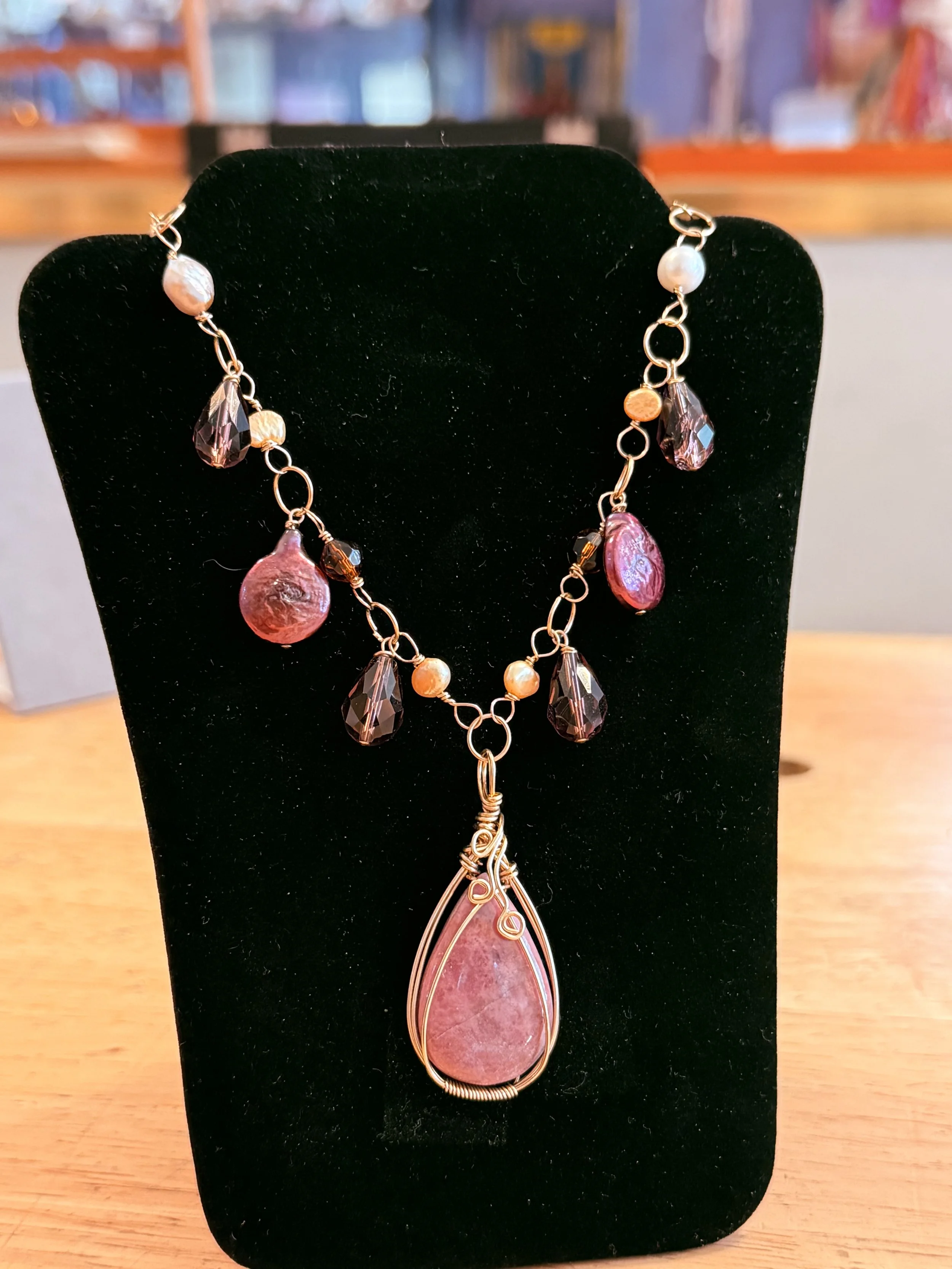 A jewelry display stand showcasing a necklace with a large pinkish stone pendant wire-wrapped with gold wire, complemented by various smaller beads and crystals in shades of pink, black, and white, arranged on a black velvet stand.