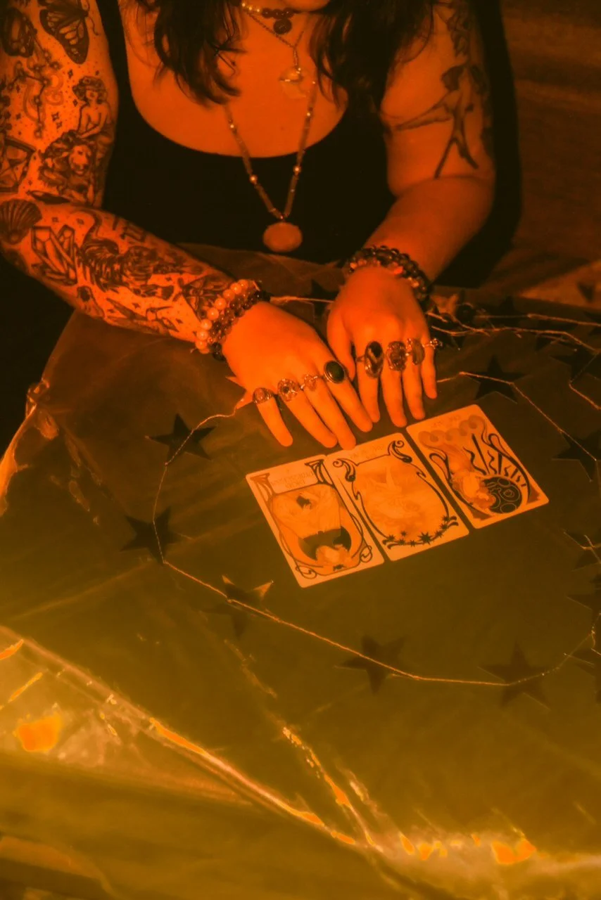 A person sitting at a table with three tarot cards laid out in front of them. The individual has tattoos on both arms, wears rings, bracelets, and a necklace, and is dressed in a black top. The scene is dimly lit with warm, orange lighting.