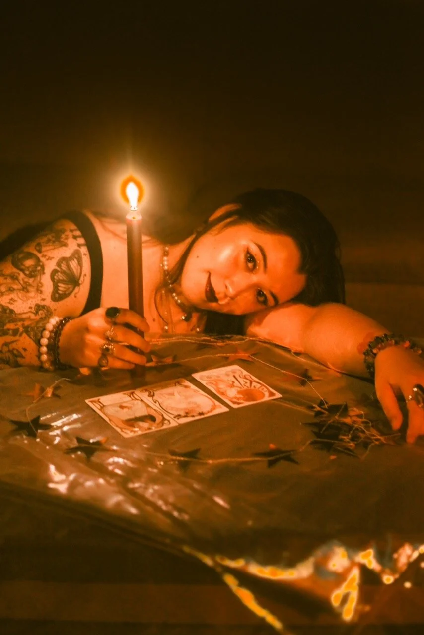A woman with tattoos and dark hair lying on a table, holding a lit candle, surrounded by tarot cards and star-shaped confetti in a dim, warm light.