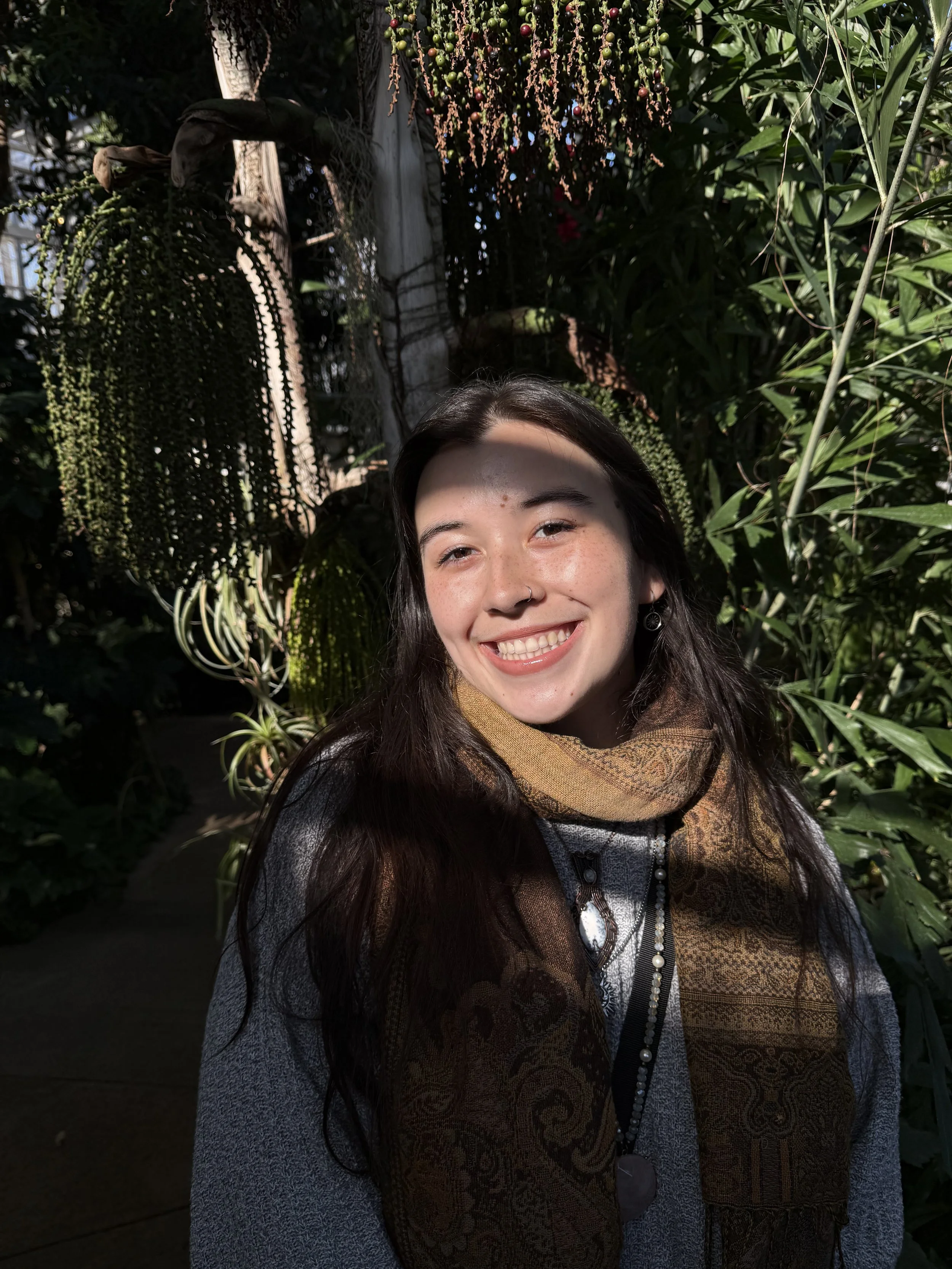 A young woman with long dark hair and a nose ring smiling in a lush, green conservatory or botanical garden with various plants and hanging foliage in the background.