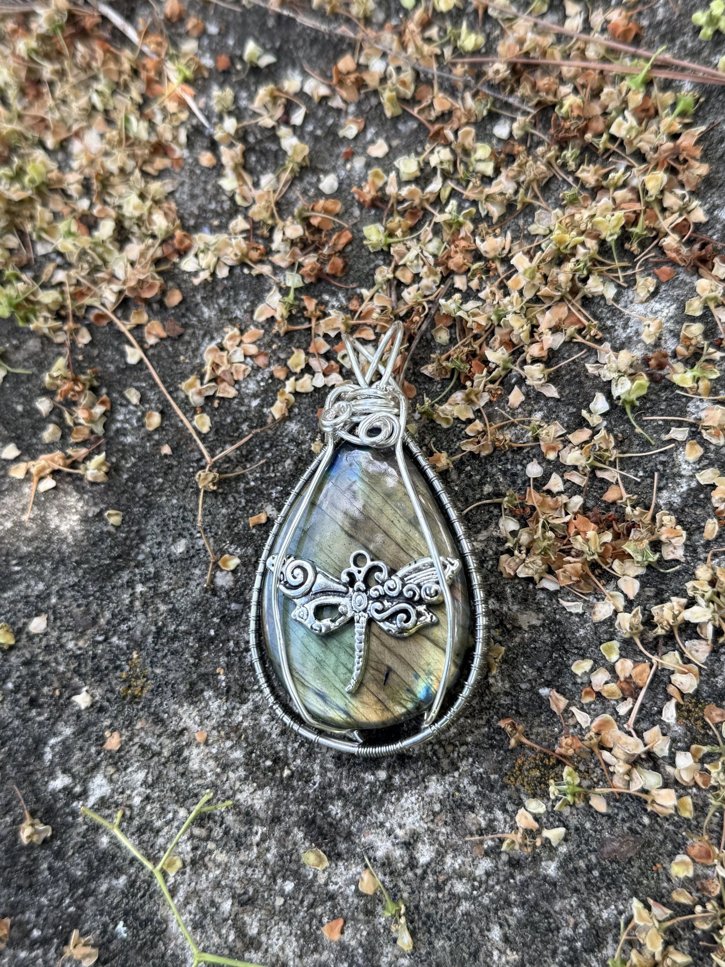 A decorative pendant necklace with an iridescent stone and silver swirled detailing, laying on a rough black surface surrounded by small, dried leaves and plant debris.