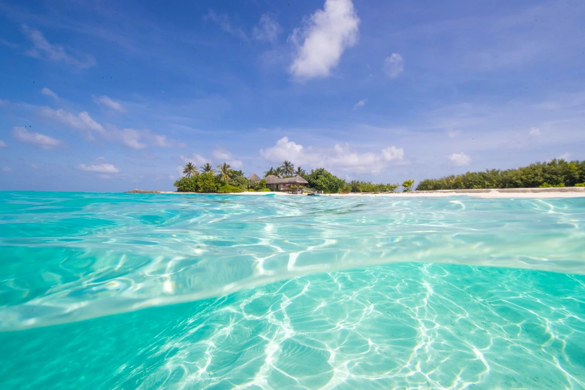 Clear turquoise water with a tropical island featuring palm trees and overwater bungalows under a partly cloudy sky.