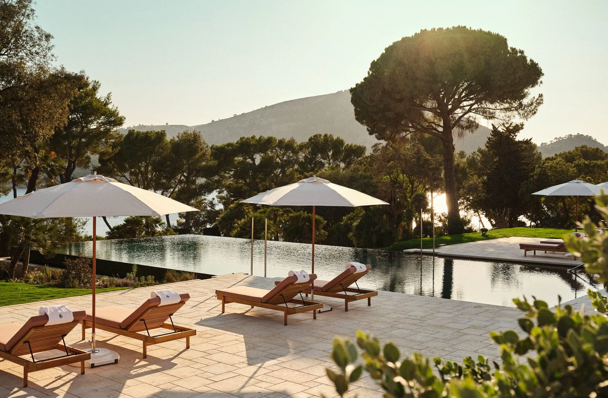Poolside area with lounge chairs, umbrellas, trees, and a mountain in the background, during late afternoon.