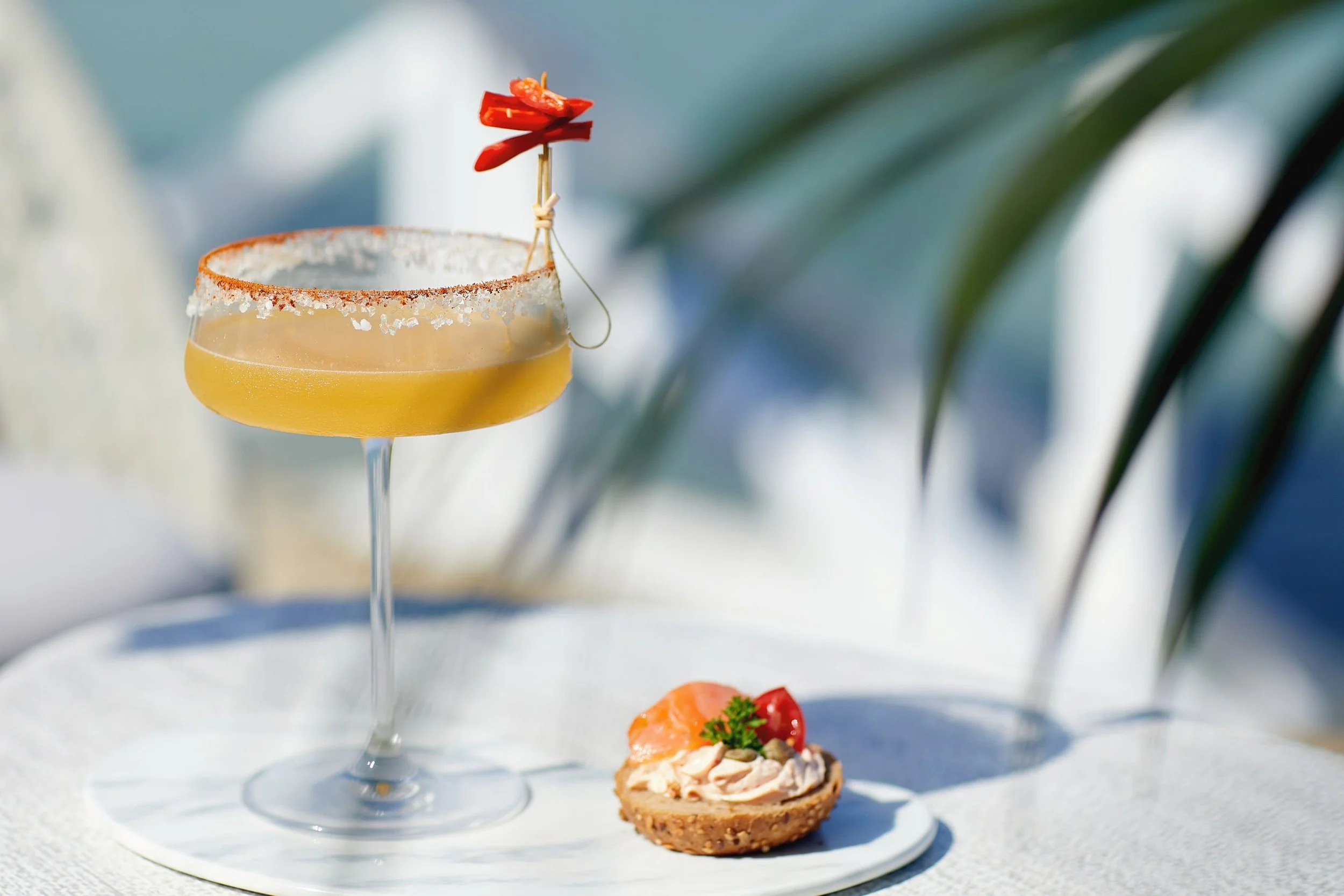 A cocktail glass with salted rim and red chili garnish, placed on a white plate with an appetizer that has a cracker topped with spread, cherry tomato, and parsley, with a blurred tropical palm background.