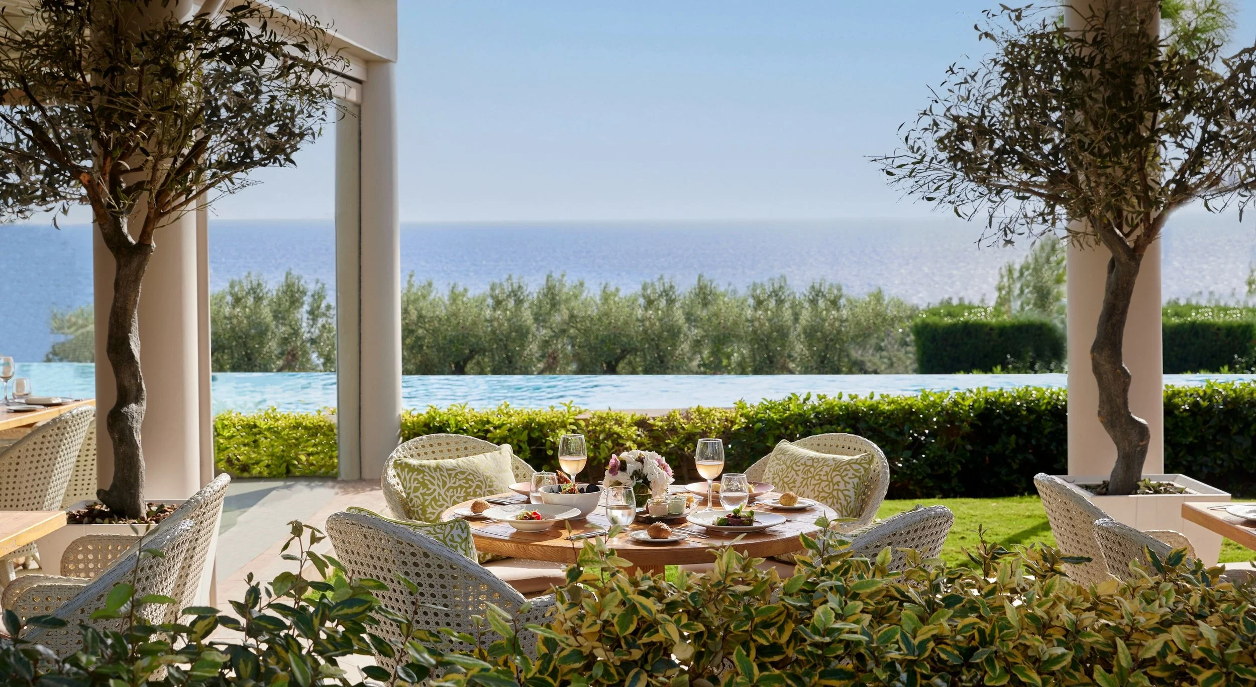Outdoor dining area with table set for a meal, surrounded by chairs, under a patio with columns, overlooking the ocean, with trees and greenery.