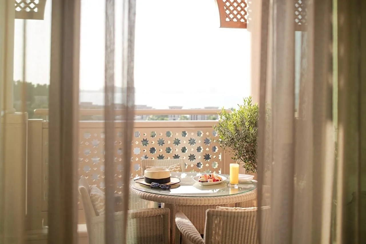 View through sheer curtains of a balcony table set with a hat, sunglasses, a pitcher of orange juice, a bowl of strawberries, and a glass of orange juice, with a patterned railing and potted plant in the background