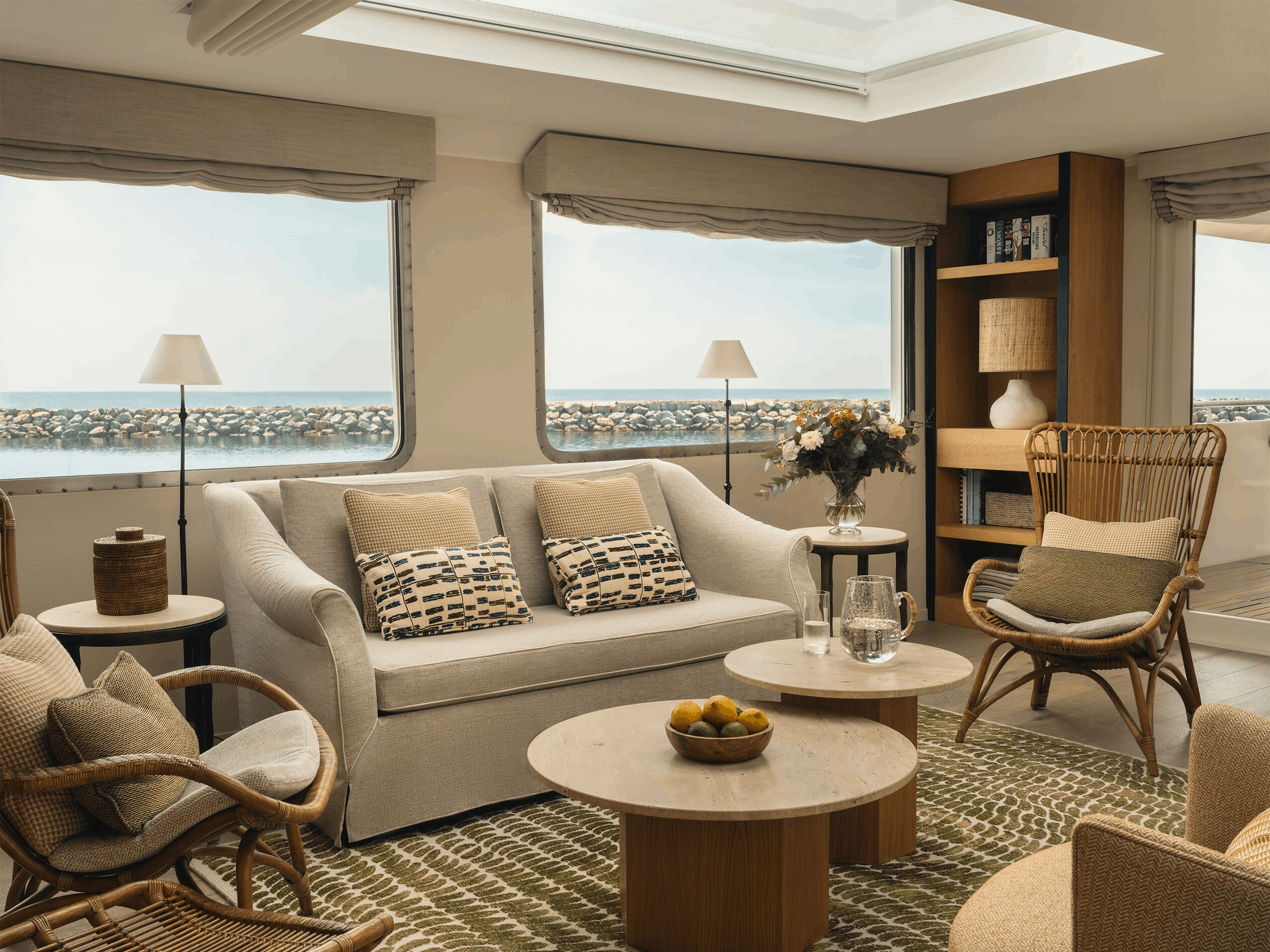Living room with large windows showing a view of the ocean and rocky shoreline. Contains a beige sofa with patterned pillows, rattan chairs, a wooden coffee table with a bowl of lemons, and floor lamps. Coastal decor with a bookshelf and vase with flowers.