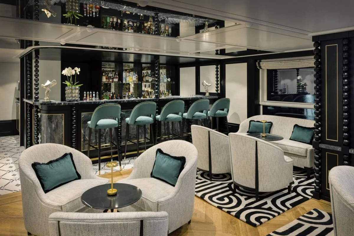 Elegant home bar area with a black and white color scheme, featuring a black marble counter with bar stools, glassware, and liquor bottles, surrounded by plush beige armchairs with teal pillows, and black and white geometric patterned rugs.