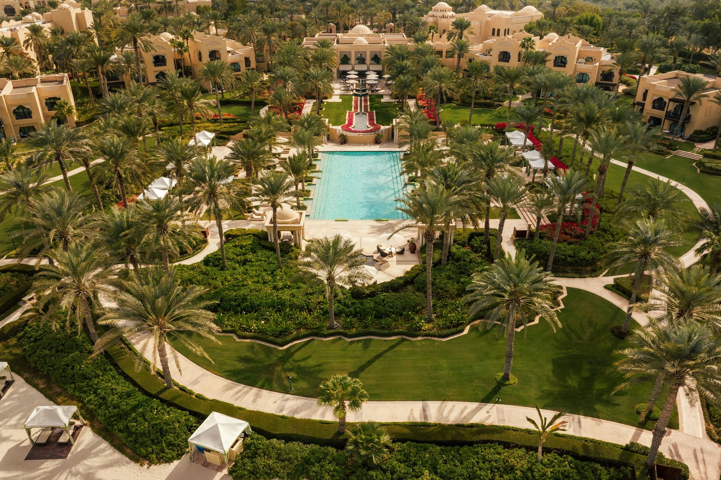 Aerial view of a luxurious resort with a central swimming pool, surrounded by palm trees, green lawns, pathways, and beige buildings.