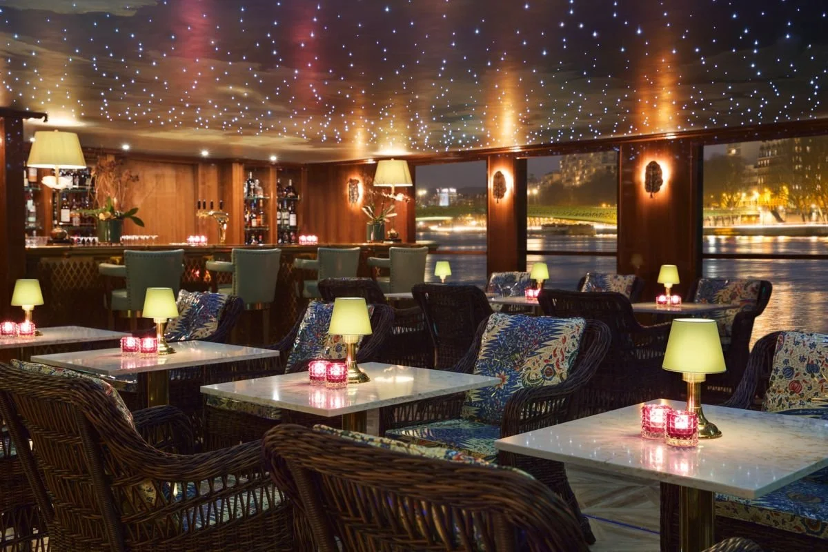Indoor restaurant with moonlit water view, decorated with star-like ceiling lights, small table lamps, and candlelit tables, featuring wicker chairs with floral cushions.