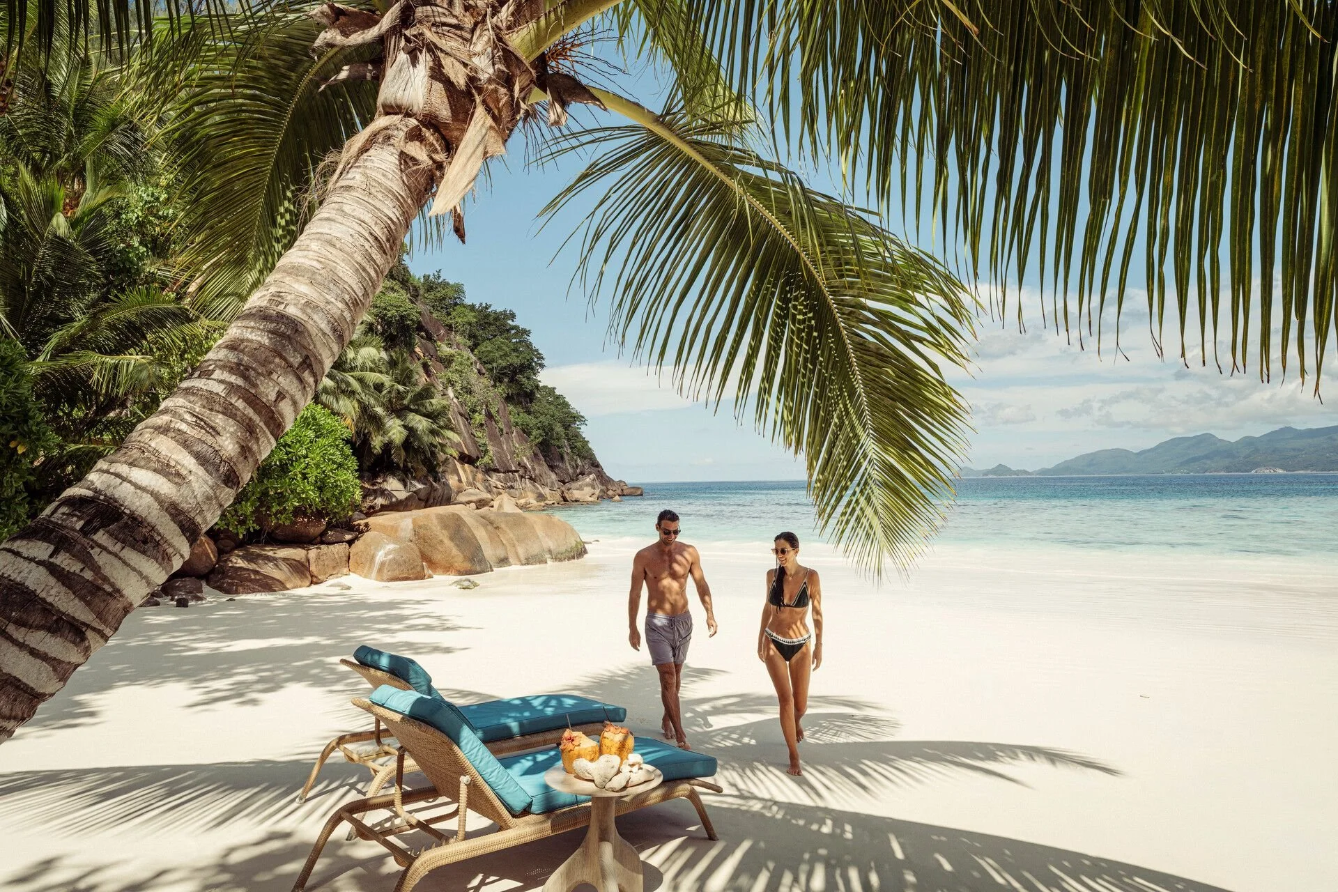 Two people walking on a tropical beach with a palm tree overhead, lounge chairs, and drinks on a table.