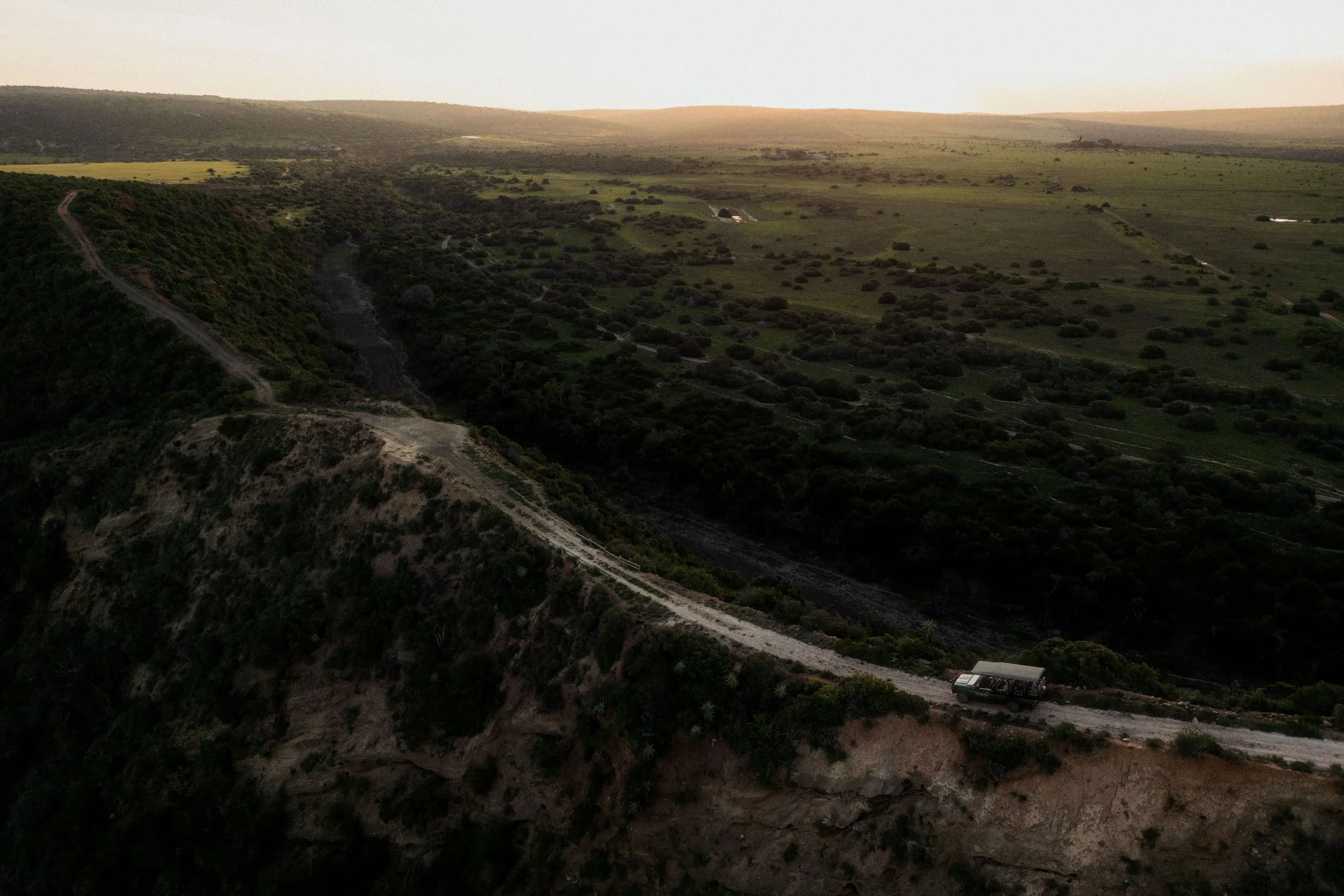 Scenic view of rolling green hills at sunset with dirt roads and a vehicle parked on one of them.