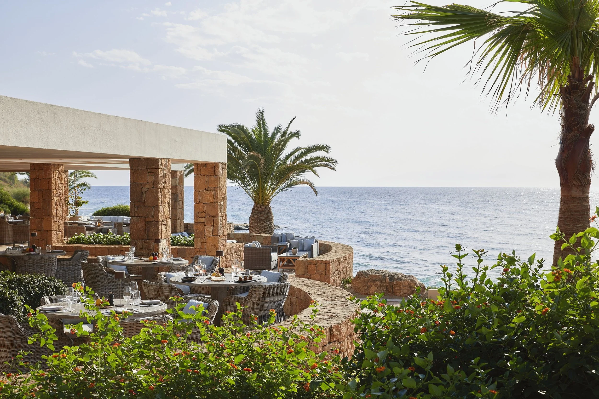 Outdoor seaside restaurant with tables set for dining, surrounded by palm trees and greenery, overlooking the ocean on a sunny day.