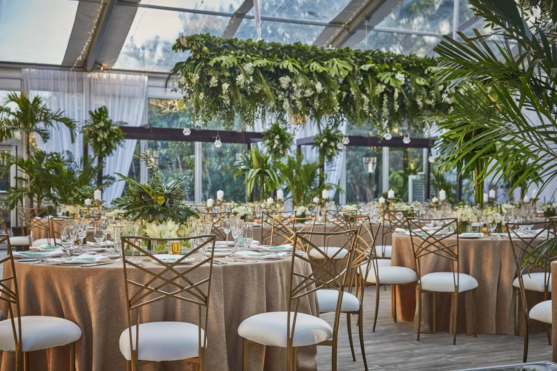 Elegant indoor event space decorated with lush green and white floral arrangements, including hanging greenery, candles, and well-arranged tables set for a formal gathering.