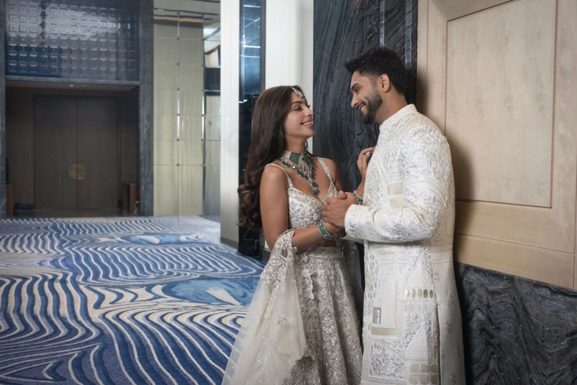 A newlywed Indian couple is standing close together in a modern hotel lobby, smiling and gazing into each other's eyes. The woman is wearing an ornate cream and silver traditional saree, adorned with jewelry and a headpiece. The man is dressed in a matching cream sherwani with intricate embroidery. The background features a patterned blue and white carpet, a black marble wall, and modern decor.