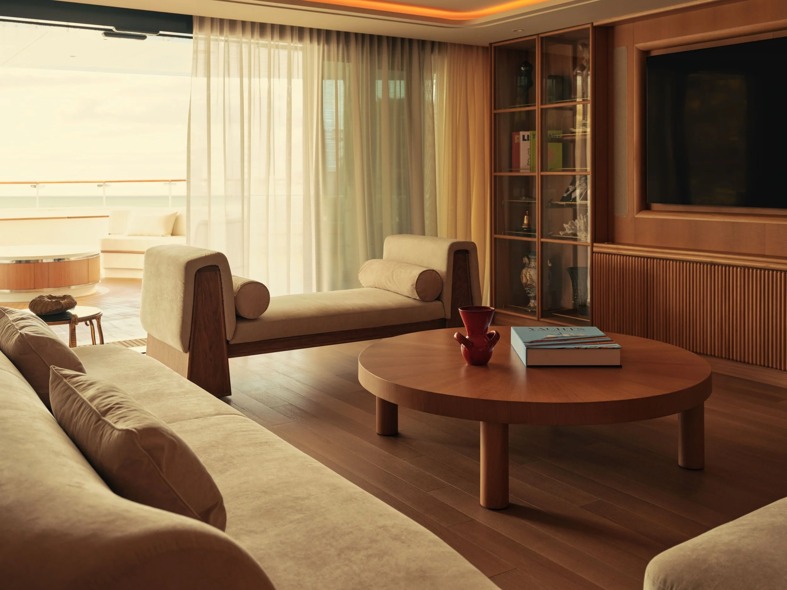 Living room with beige sofas, a round coffee table with books and a red vase, and large window with sheer curtains letting in natural light. There is a wooden cabinet with glass doors and a large flat-screen TV on the wall.