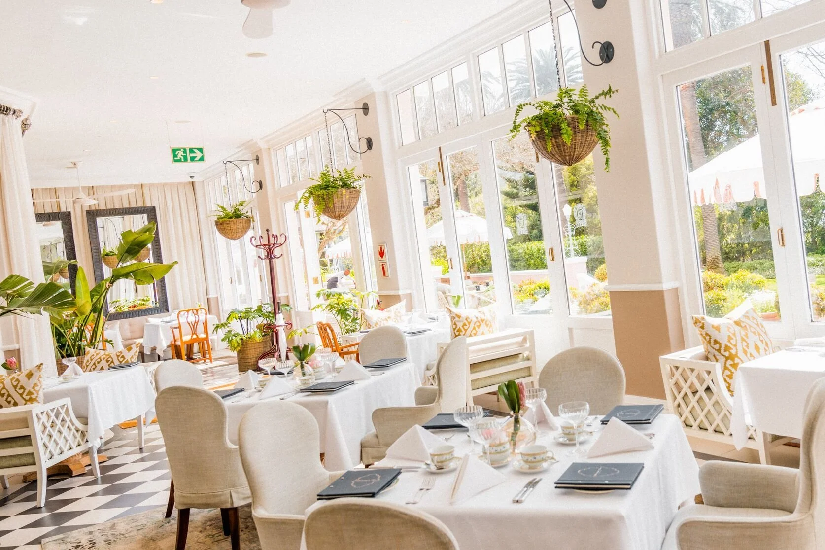 Brightly lit restaurant interior with white walls, large windows, and hanging fern plants, set for dining with white tablecloths, napkins, glasses, and floral centerpieces.