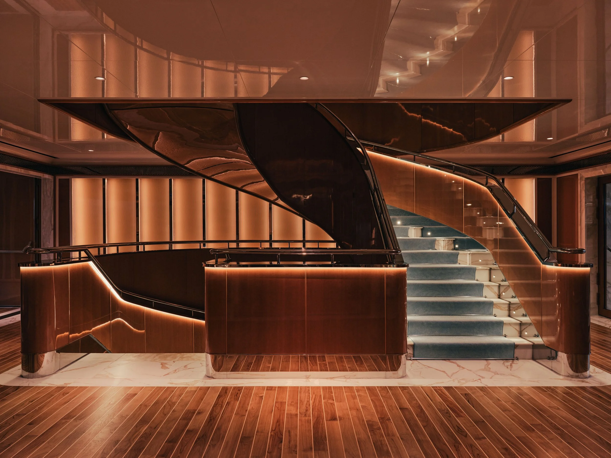 Interior of a modern building featuring a curved staircase with glass railing, illuminated by soft lighting, with wooden paneling and marble flooring.