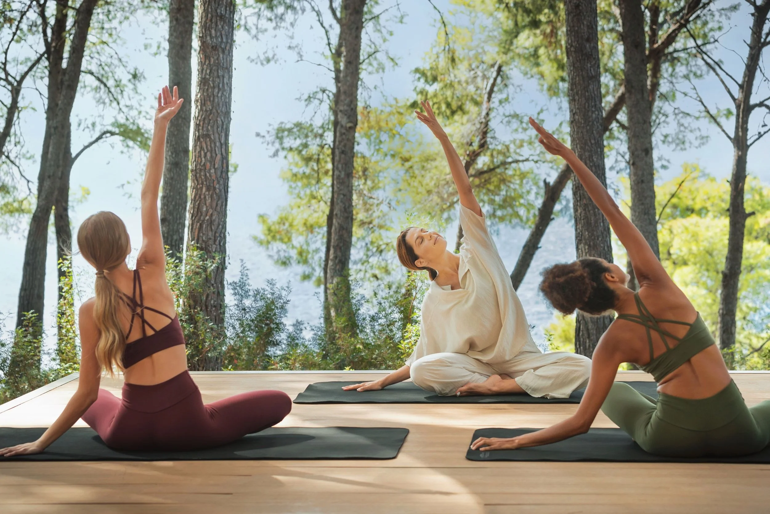 Group of people practicing yoga outdoors in a forest with tall trees, sitting on yoga mats with sunlight filtering through the leaves.