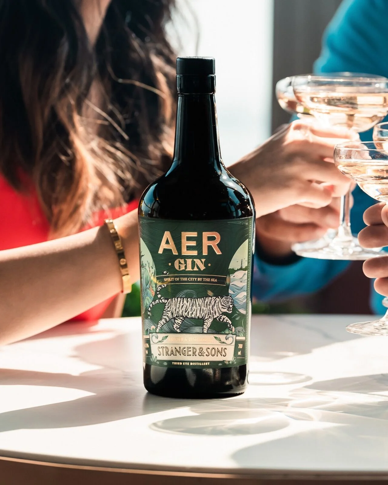 A bottle of AER GIN on a white table with people in the background holding cocktail glasses.