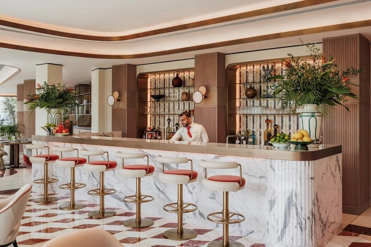 Elegant bar interior with marble counter, high stools with red and white cushions, floral arrangements, and a bartender preparing drinks in a sophisticated setting.
