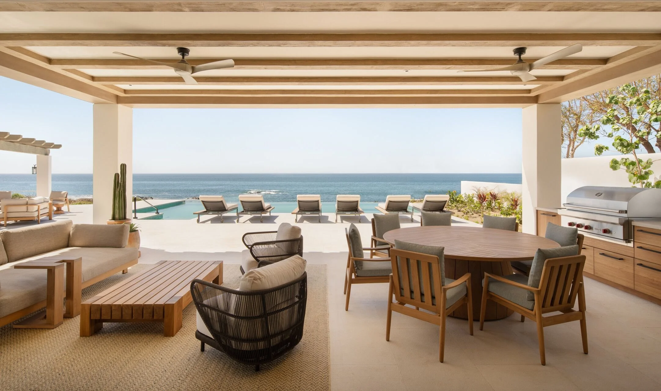 Indoor living space with a view of a swimming pool and ocean, furnished with sofas, chairs, a dining table, and an outdoor kitchen grill, under a wooden ceiling with ceiling fans.