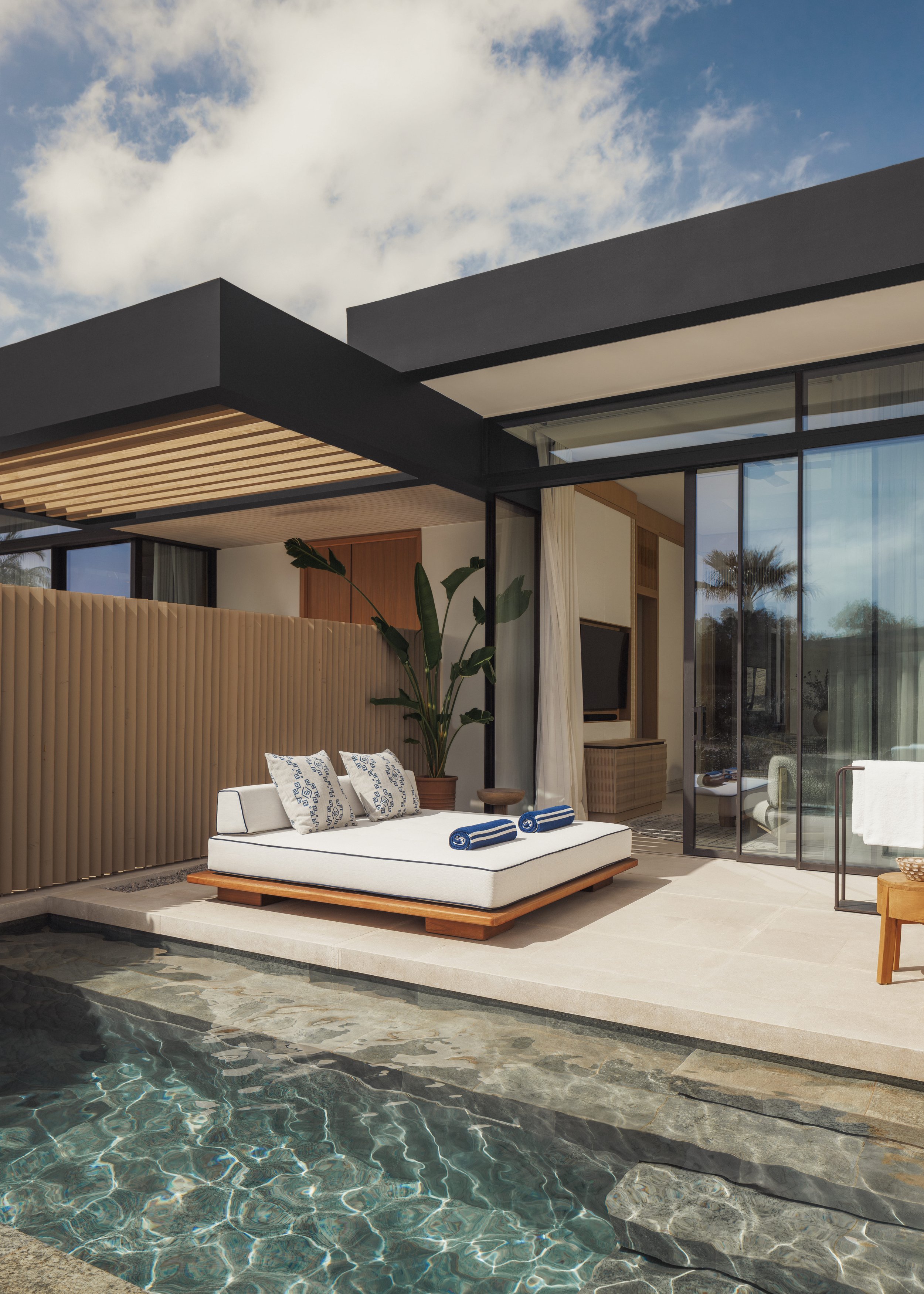 Modern outdoor patio with a poolside seating area, including a white cushioned lounge with blue and white throw pillows, a wooden side table, and surrounding plants, with the house featuring large glass windows and black framing, under a partly cloudy sky.