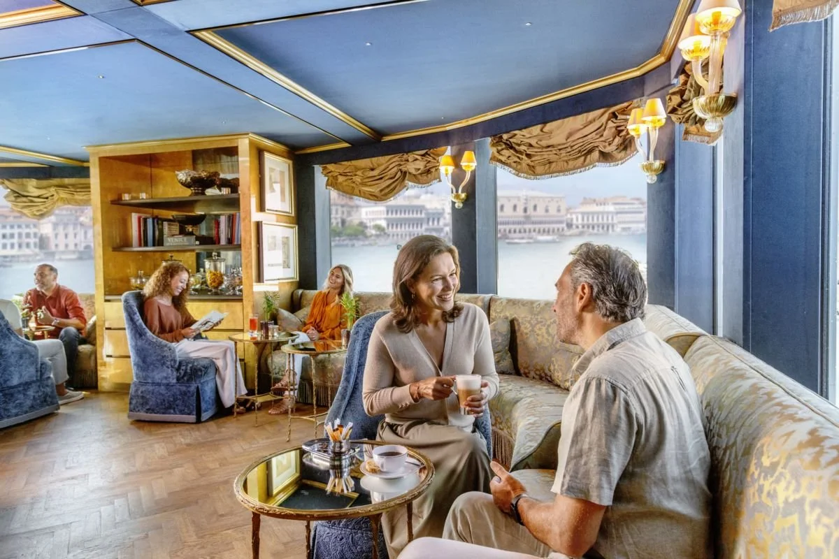 People relaxing and enjoying drinks in a bright, upscale cafe or lounge with large windows overlooking a river and historic buildings.