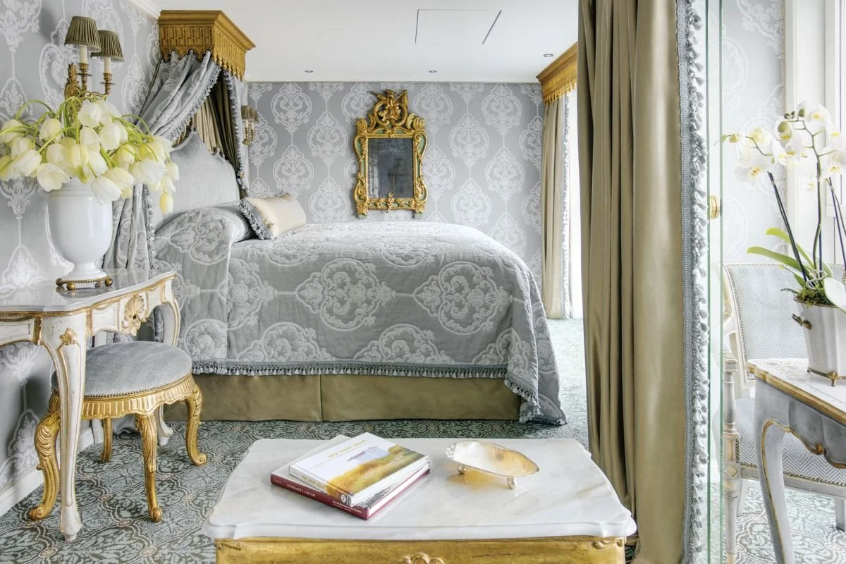 Luxurious bedroom with a white and gold color scheme, ornate furniture, a large bed with patterned quilt, floral wallpaper, and elegant drapes.