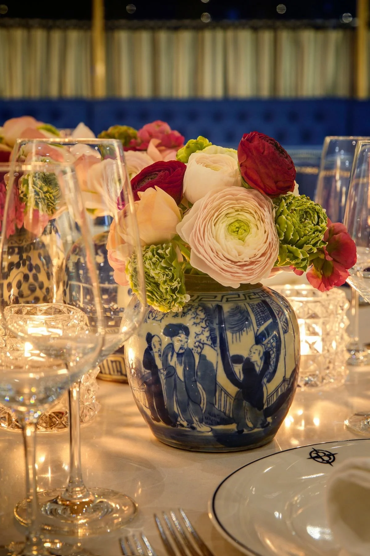 Elegant table setting with a blue and white floral patterned vase holding a bouquet of pink, white, red, and green flowers, surrounded by crystal glasses, a plate with a decorative border, and silver utensils, with a background of bookshelves.