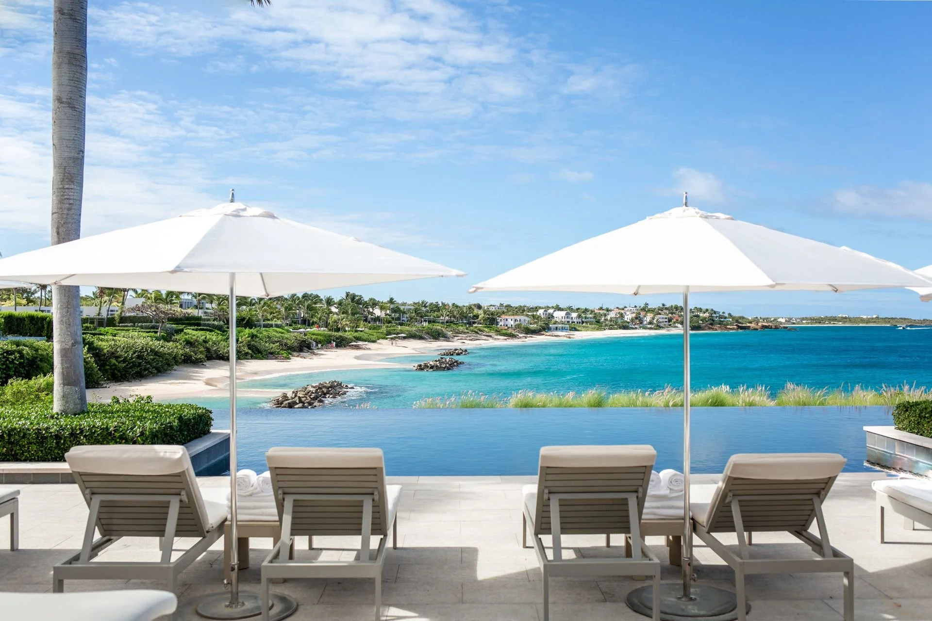 Poolside view with lounge chairs and white umbrellas overlooking a tropical beach and ocean with greenery and houses in the background.