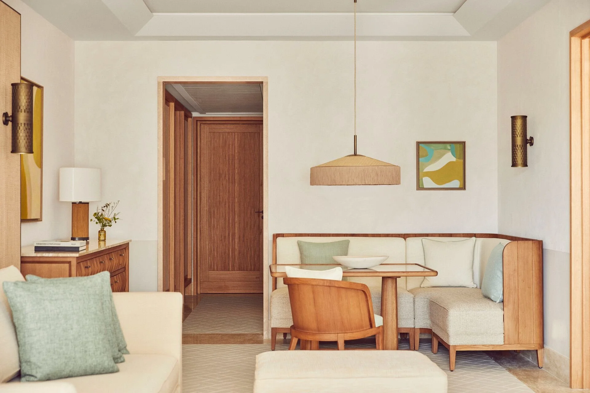 A modern living room with white and green cushions on a cream sofa, a wooden dining table with chairs, a wall-mounted light fixture, framed abstract art, and a wooden door.