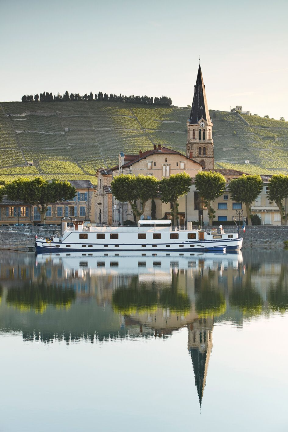 A scenic view of a European town with a church steeple, colorful buildings, and a boat on calm water reflecting the town and a hillside with vineyards in the background.