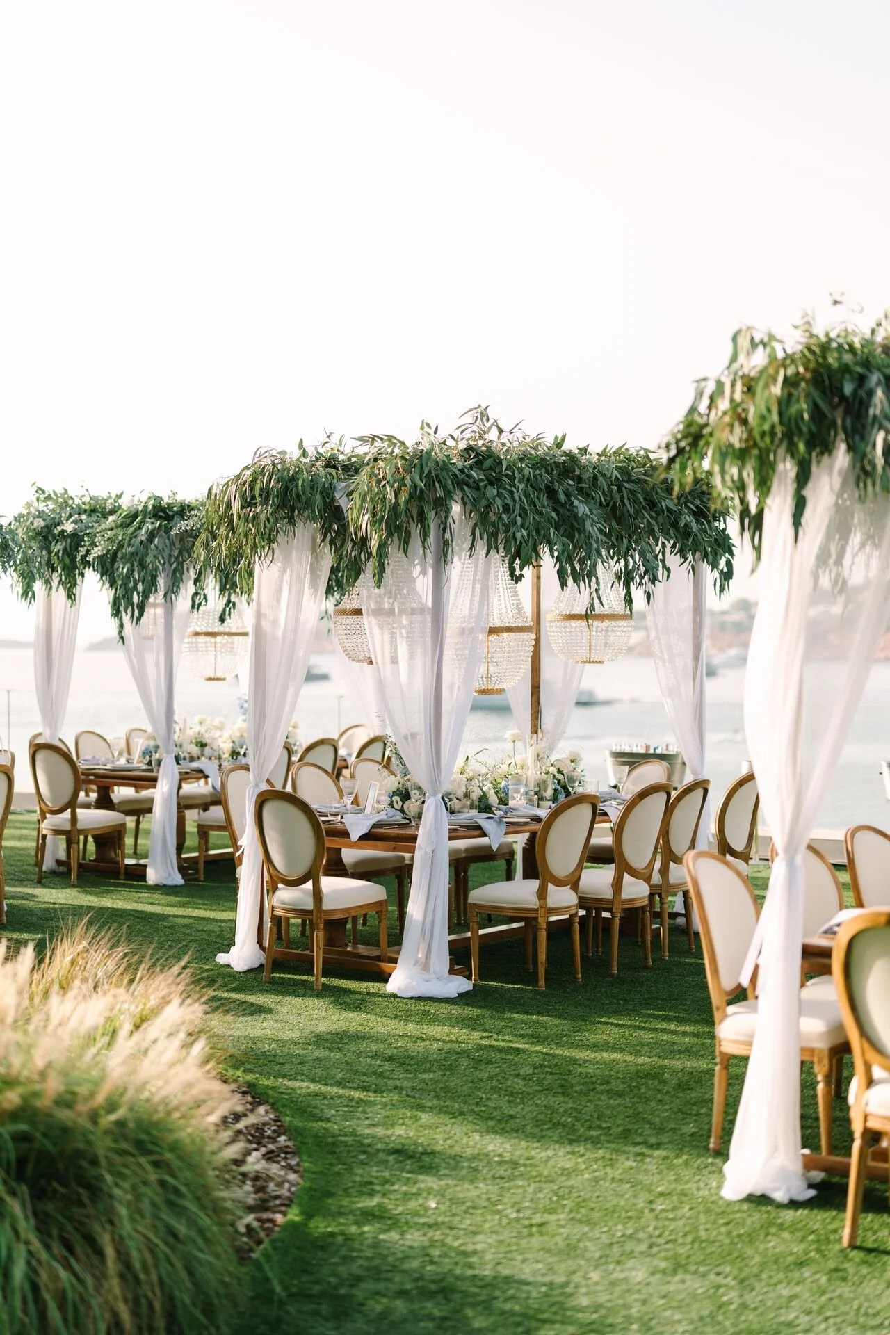 Elegant outdoor wedding setup with round tables surrounded by chairs on a grassy area, decorated with white drapery and lush green foliage overhead, overlooking a body of water.
