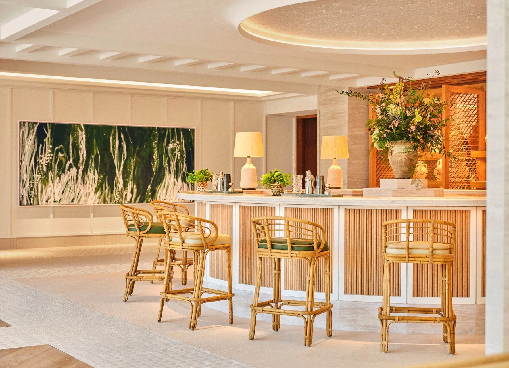 Elegant hotel lobby with a reception area featuring a beige marble counter, wooden accents, green plants, yellow lamps, and a large floral arrangement in a vase. Pastel-colored chairs with wooden frames are arranged in front of the counter, and a large abstract painting decorates the wall in the background.