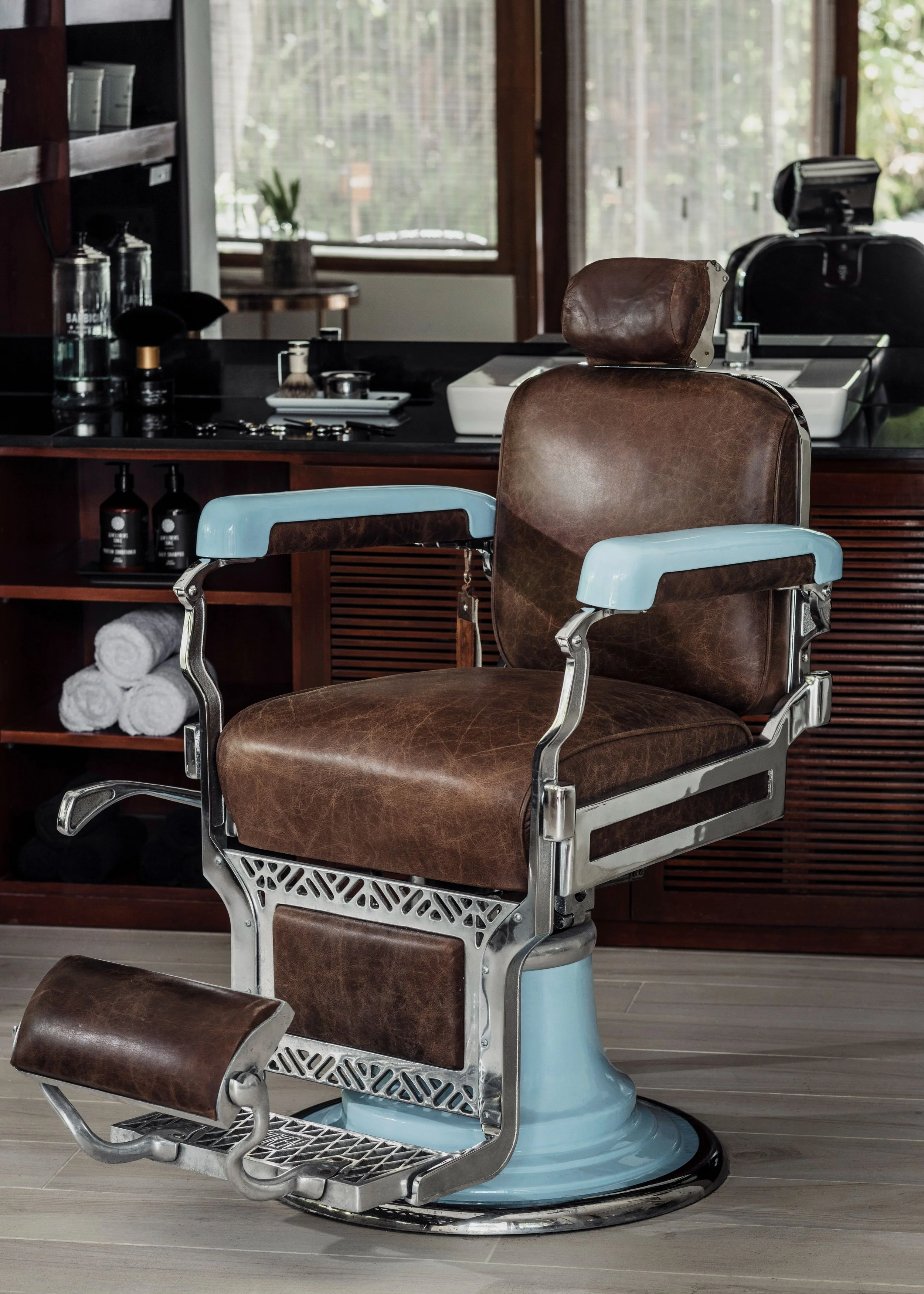 Vintage barber chair with brown leather upholstery and light blue metal accents, in a barber shop with large windows and wooden furnishings.