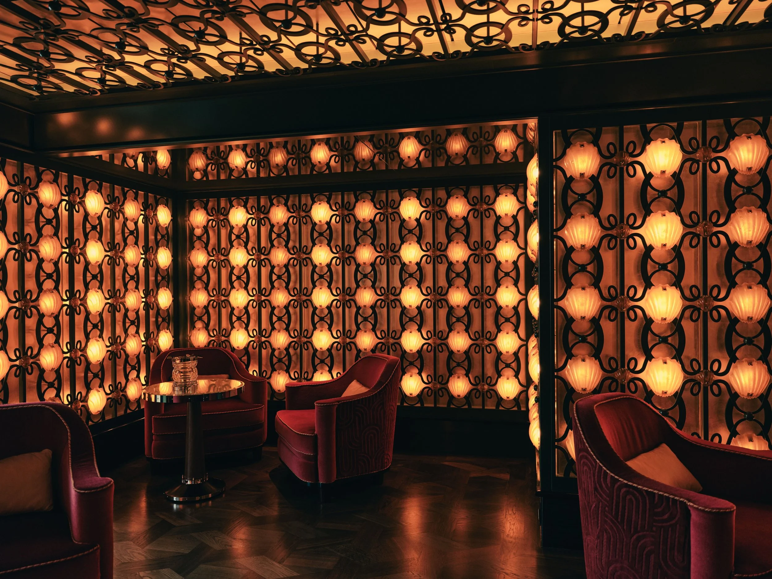 Interior of a room with warm lighting, featuring a decorative wall with illuminated lanterns and intricate black wrought iron patterns, velvet chairs, and small tables.
