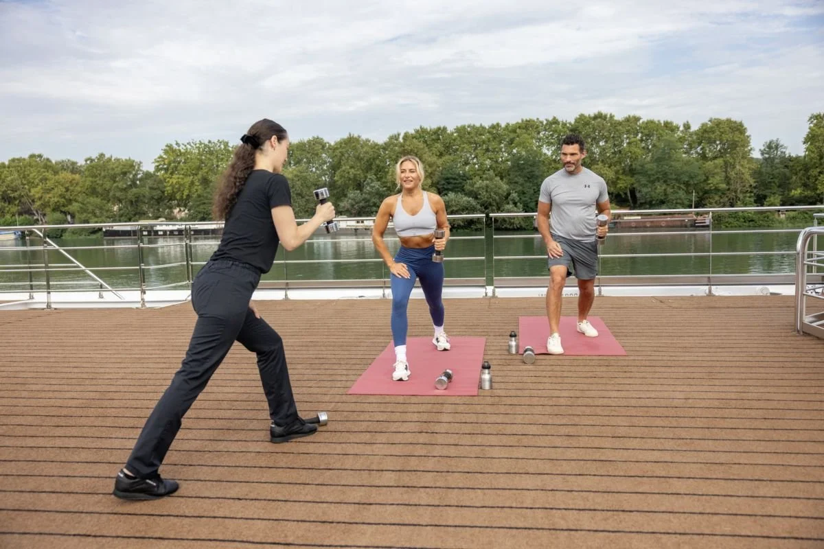 A woman and a man exercising on a deck by the water, holding water bottles and doing lunges, while a trainer records them with a camera.