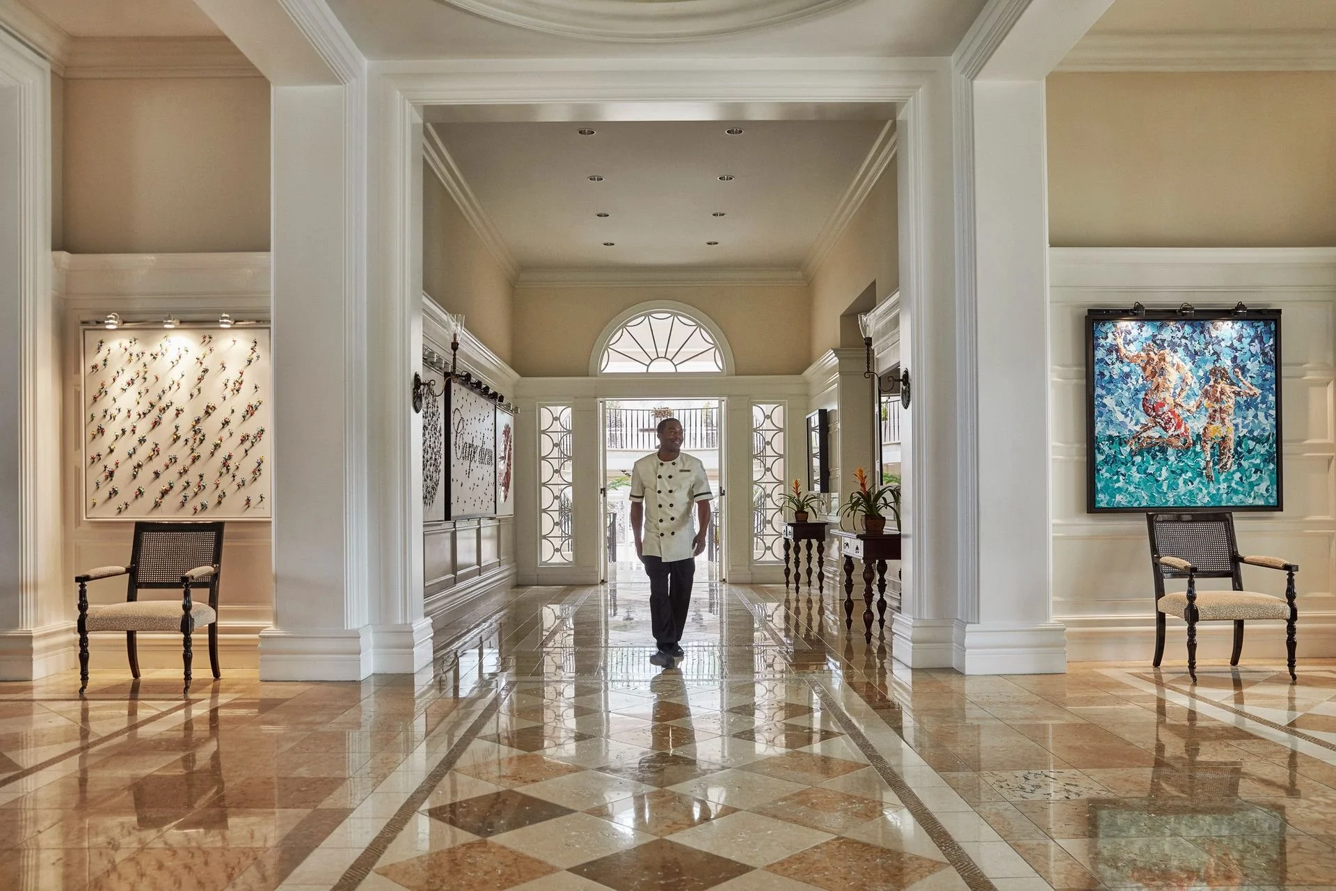Elegant hotel lobby with marble floor, white walls, and decorative molding. A man in a chef's uniform walks toward the entrance. Artworks and chairs are visible on the walls.