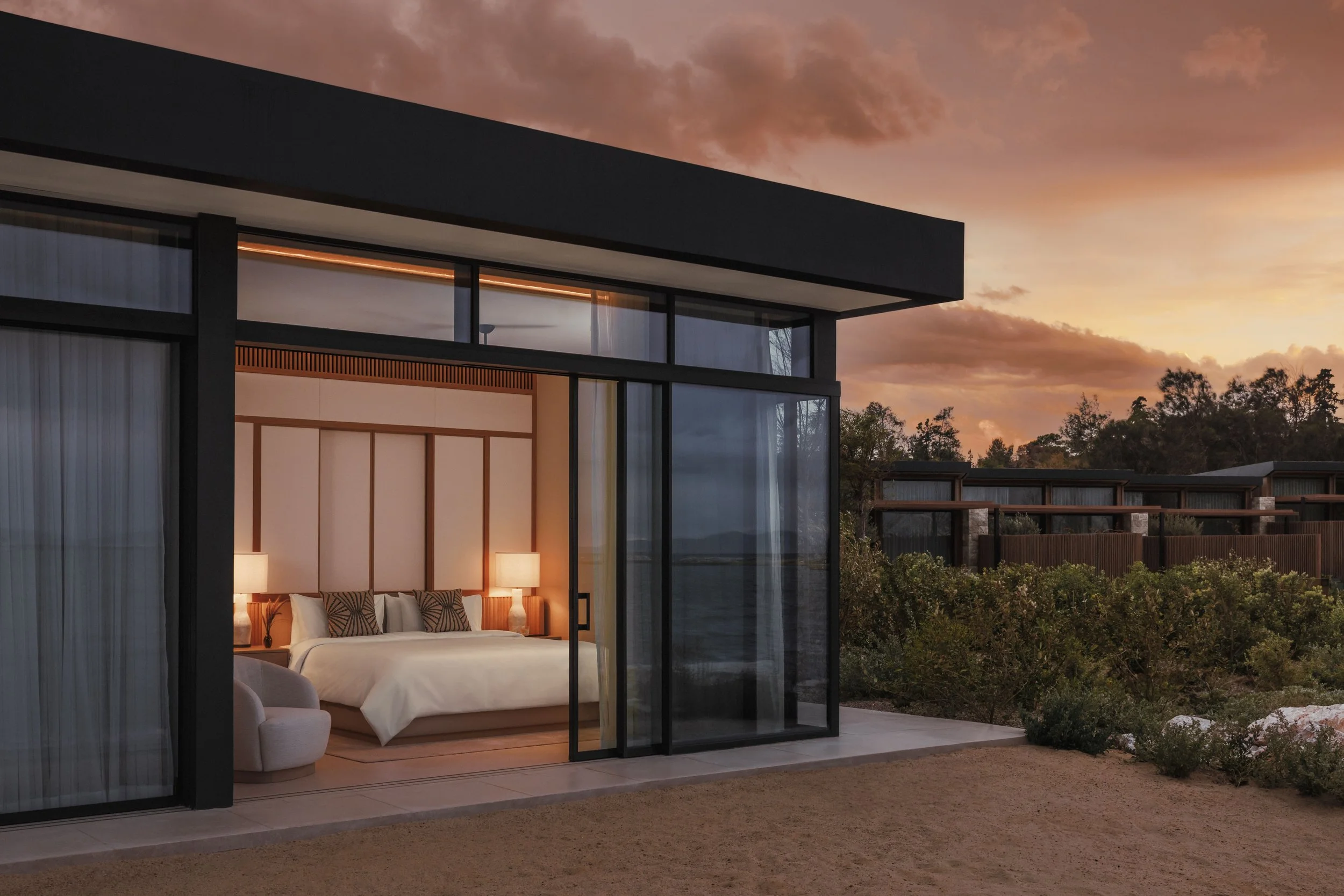 Modern bedroom inside a house with large glass sliding doors open to the outdoor sunset, showing a bedroom with white bedding, lamps, and decorative pillows.